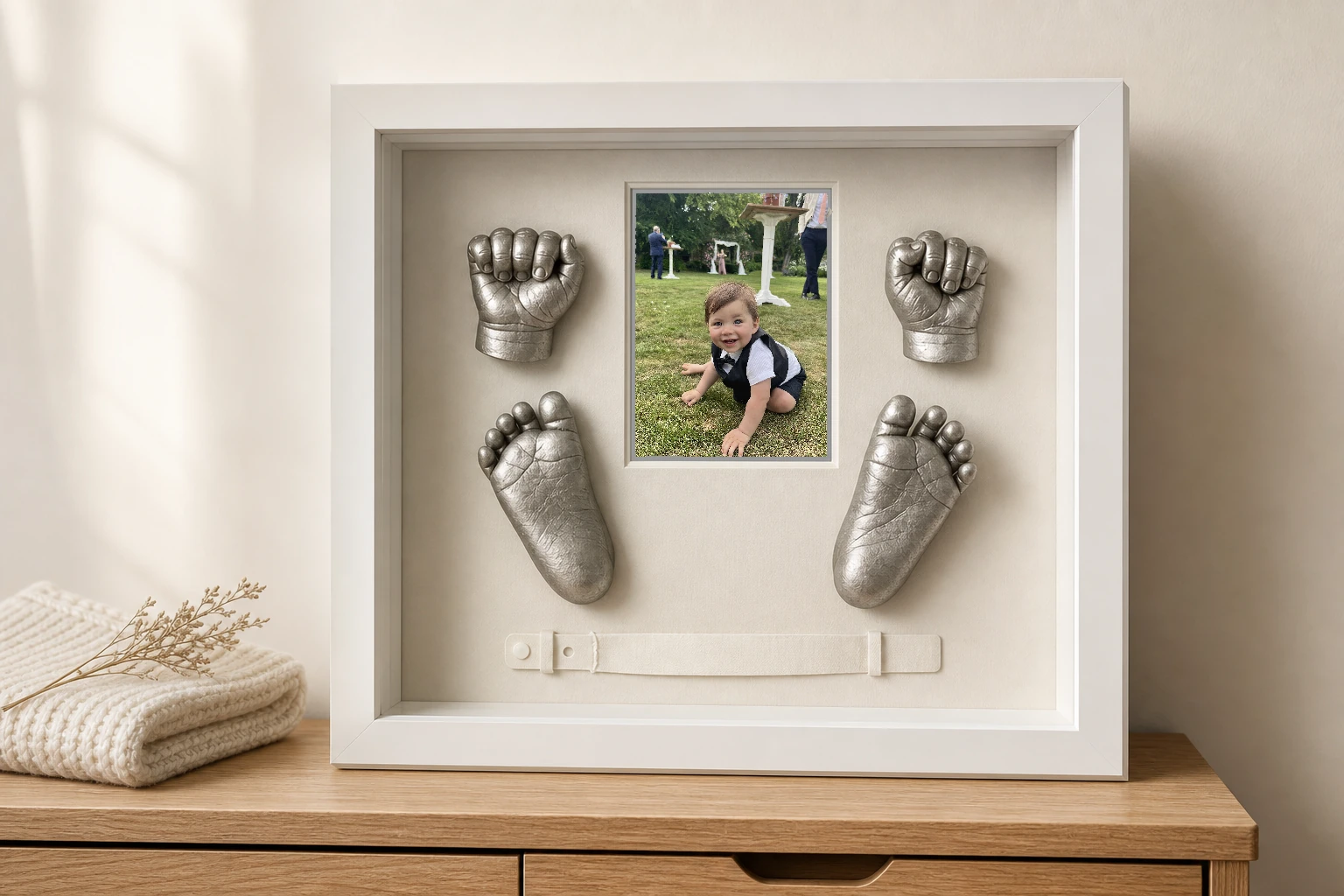 White nursery shadow box displaying silver baby hand and foot castings, a smiling baby portrait, and a hospital band